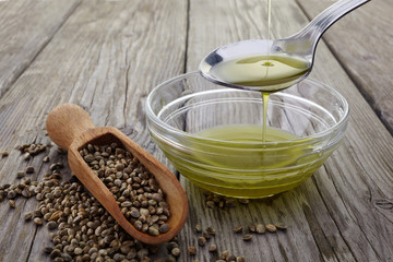 Pouring hemp oil into glass bowl and hemp seeds on wooden background