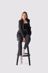 Fototapeta premium Full length portrait of beautiful young girl sitting on tall chair in black jeans and jacket over gray background
