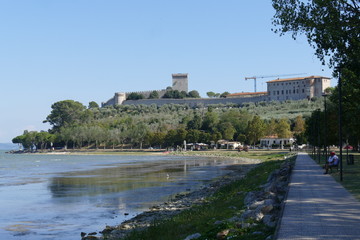 Fototapeta na wymiar Castiglione del Lago walk on the green lakeside of Trasimeno lake with the castle on the hill in the background