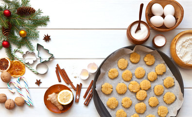 Sweet christmas cookies on baking sheet before baking. Bowl with flour, eggs, nuts, ginger, cinnamon, lemon, sugar and decorated Christmas tree branches on white table. Flat lay, copy space, close-up