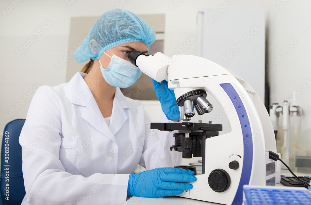 Poster young laboratory assistant in mask looking through modern microscope