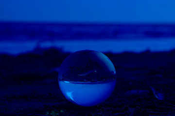 Glass ball in focus on the beach send near the sea with upsidedown reflection and lighs flecks on the defocused background. Horizontal with copy space. Classic, blue monochrome, trend 2020