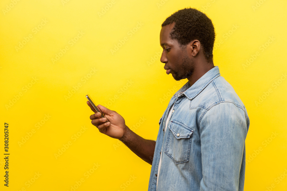 Wall mural side view of serious man in denim shirt using cellphone, typing text message or dialing number with 