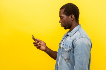 Side view of serious man in denim shirt using cellphone, typing text message or dialing number with calm attentive expression, technology concept. indoor studio shot isolated on yellow background