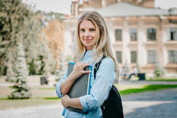 A blonde student girl is smiling and holding a folder and a notebook in her hands at university background. Girl is taking exams at university