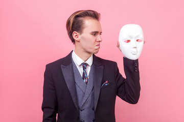 Profile of upset depressed man in elegant suit with stylish haircut looking at white mask with pursed lips desperate face, hating his hidden personality. studio shot isolated on pink background