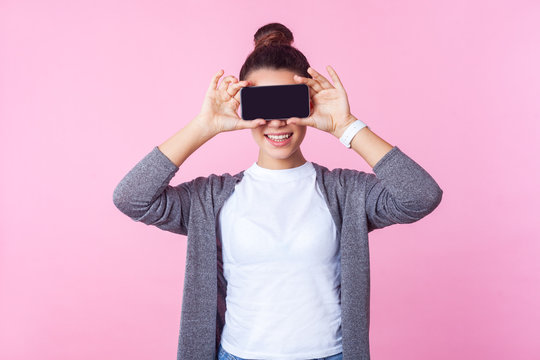 Portrait of brunette teenage girl with bun hairstyle in casual clothes smiling and covering eyes with cell phone, hiding face to be anonymous in internet. indoor studio shot isolated, pink background