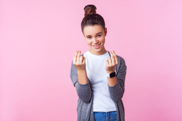 Portrait of sly brunette girl with bun hairstyle in casual clothes showing money gesture and looking cunning at camera, teenager demanding more allowance. studio shot isolated on pink background
