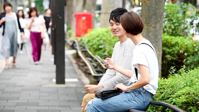 歩道の脇のベンチでスマートフォンを見ながら会話するカップル