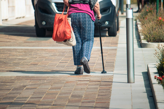 walking stick detail, an old female person with bags walks using a support for deambulation
