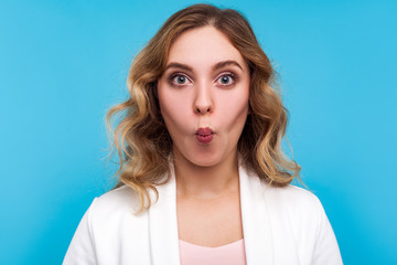 Naklejka premium Portrait of funny beautiful woman with wavy hair in white jacket making fish face with pout lips and amazed big eyes, surprised goofy expression. indoor studio shot isolated on blue background