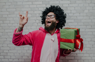 Merry Christmas and happy New Year! Cheerful bearded young man with curly hair in pink dragon pajamas with Christmas gift box. Crazy emotions. Pajamas party