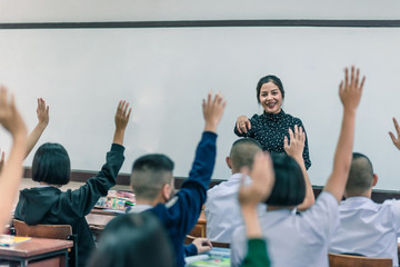 A smiling Asian female high school teacher teaches the white uniform students in the classroom by asking questions and then the students raise their hands for answers.