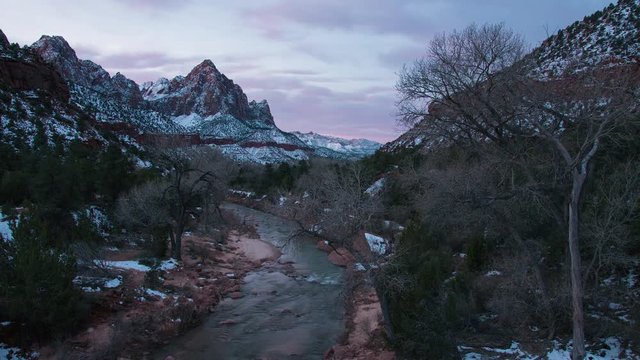 Timelapse of colorful sunrise from the Zion bridge as the sky lights up looking down the Virgin River towards The Watchman peak covered in snow in winter.