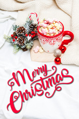Red mug with marshmallows and winter ornaments on a white sheets with text Merry Christmas