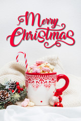 Red mug with marshmallows and winter ornaments on a white sheets with text Merry Christmas