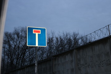 A road sign marking a dead end against a stone wall with barbed wire and the evening sky. Photo at...