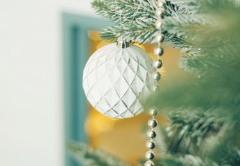 White Christmas ball toy and a silver beads on the snow branch of a green tree. Festive yellow lights from a house window. Christmas winter concept. Top view