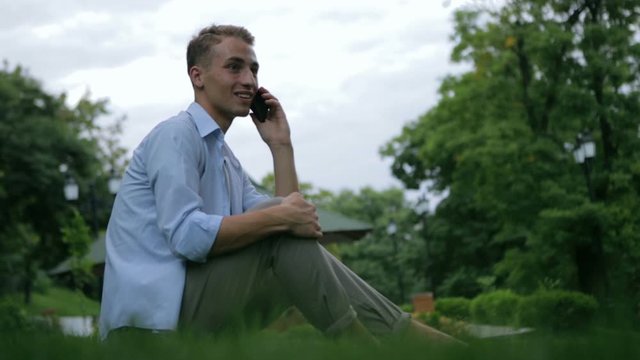 handsome guy sits barefoot on grass and talks on smartphone smiling against green plants in local park closeup