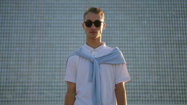 serious blond guy in sunglasses poses standing in front of glass urban building wall in city center close view