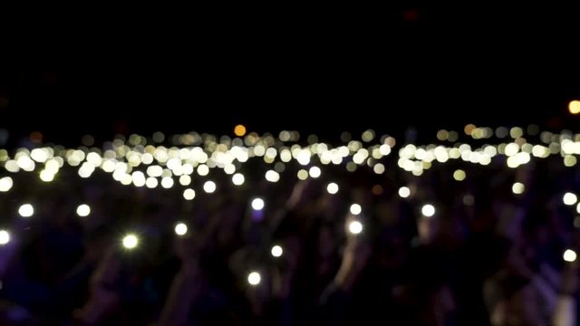 Defocused spectators in hall with lights waving in the dark. Action. Audience holding phones with shining flashlights during the concert, romantic atmosphere.
