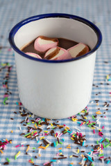 Close-up of cup of rustic chocolate and marshmallows, with unfocused close-up, on blue and white checkered tablecloth with chocolate and color vermicelli, vertically, with copy space