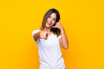 Young woman over isolated yellow background making phone gesture and pointing front