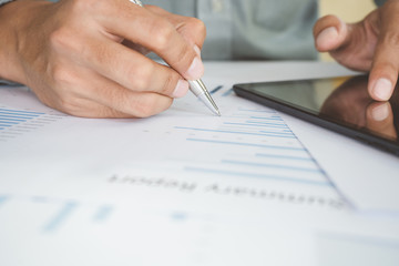 Asian businessman is using a pen to write a document report with laptop, tablet on a desk in the office. Staff checking the paper and planning the company's finances