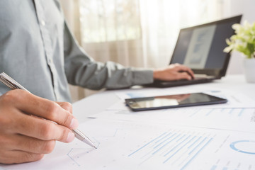 Asian businessman is using a pen to write a document report with laptop, tablet on a desk in the office. Staff checking the paper and planning the company's finances