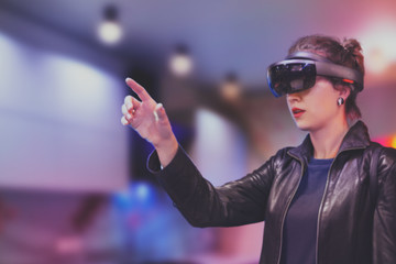 Portrait of young Caucasian woman using augmented and virtual reality with holographic hololens glasses. Pink, magenta, blue blurred background. Future technology concept. 