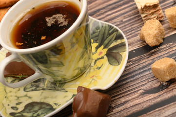 A Cup of black tea, tea leaves, pieces of brown sugar, oatmeal cookies, waffles, pieces of chocolate on a wooden background. Close up.