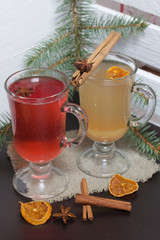 Glasses with a drink of orange and red are on a piece of linen. Slices of dried orange and anise float in them. Nearby are cinnamon sticks and dried oranges and a spruce branch.