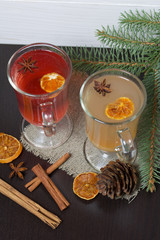 Glasses with a drink of orange and red are on a piece of linen. Slices of dried orange and anise float in them. Nearby are cinnamon sticks and dried oranges and a spruce branch.