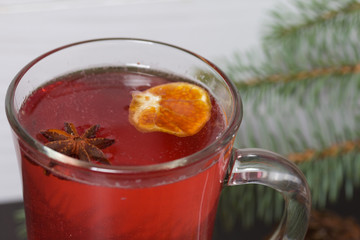 A glass of red drink stands on a piece of linen. A slice of dried orange and anise floats in it. Nearby are cinnamon sticks and dried oranges and a spruce branch.