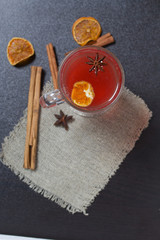 A glass of red drink stands on a piece of linen. A slice of dried orange and anise floats in it. Nearby are cinnamon sticks and dried oranges.