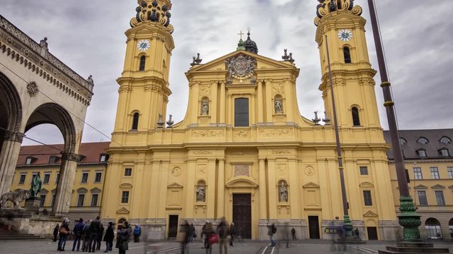 Time-lapse of a moody sky in the background with people walking in the foreground, in front of the yellow church of Odeonsplatz in Munich, Germany.