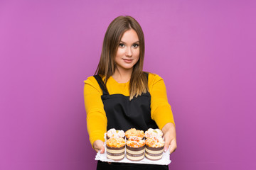 Teenager girl holding lots of different mini cakes over isolated purple background