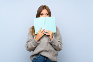Teenager girl with sweater over isolated blue background holding and reading a book