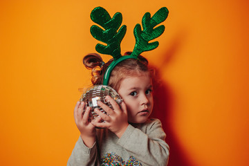 Christmas card with copy space. Little kid with green christmas antlers on head. Baby girl holds disco ball in hands on the orange background. Baby girl deer.