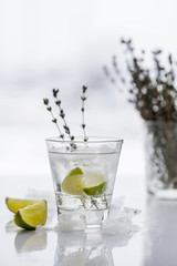 Glass with cocktail with lime, thyme and ice on a light background with multi-colored lights.