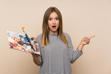 Teenager girl holding a palette over isolated background surprised and pointing finger to the side
