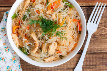 Funchosa with vegetables and chicken on a white plate on a wooden table, selective focus