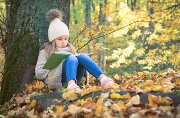 Cute little girl sitting with a book on her lap under the tree in public park. Girl sitting thoughtfully on the ground covered with yellow leaves in the autumn season sunny weather.