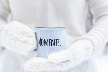female hands in white mittens holding a blue mug with the inscription MOMENTS