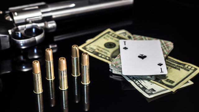 Dangerous gambling for money. A pistol with cartridges, money dollars and a deck of playing cards with an ACE of spades lie on a glass table. Dangerous gambling debts and loans.