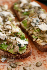 Two large sandwiches made from black gluten-free bread, avocado, cheese, and pumpkin seeds.