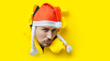 A young guy with a beard and a mustache with duck lips in a Santa Claus or Snow Maiden hat with white pigtails peeps out of a torn hole in yellow paper. The concept of Christmas, New Year. Copy space.