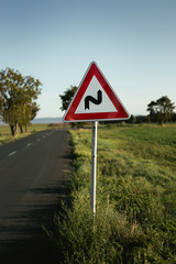 Photo of illuminated and warning road sign - double curve next to the road on meadow. Double Bend warning road sign on country road with trees and beautiful landscape on background - European sign.