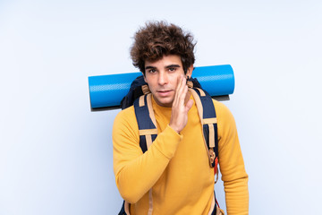 Young mountaineer man with a big backpack over isolated blue background whispering something