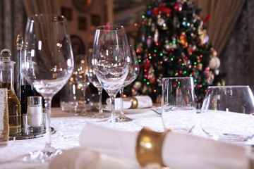 Table setting for the New year. The Christmas tree is out of focus. Lots of empty glass glasses on the table. Photo in the interior.